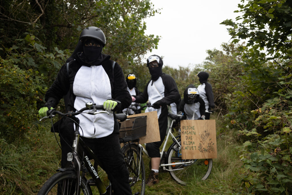 Mehrere Menschen in Pinguinkostümen auf Fahrrädern auf einem Feldweg zwischen Gebüsch und Bäumen. An einem Fahrrad ist ein Holzschild "Reiche machen das Klima kaputt" mit Dagobert Duck, der in Geldscheine springt.